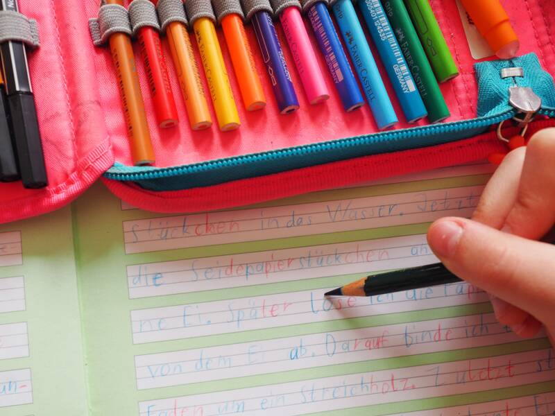 Eine Kinderhand hält einen Buntstift mit blauer Mine und schreibt in ein Schreibheft mit vorgegebenen Linien in einem weißen Schriftfeld Wörter. Der Rest des Heftes ist hellgrün und auf dem Heft liegt ein offenes Federmäppchen mit bunten Buntstiften.
