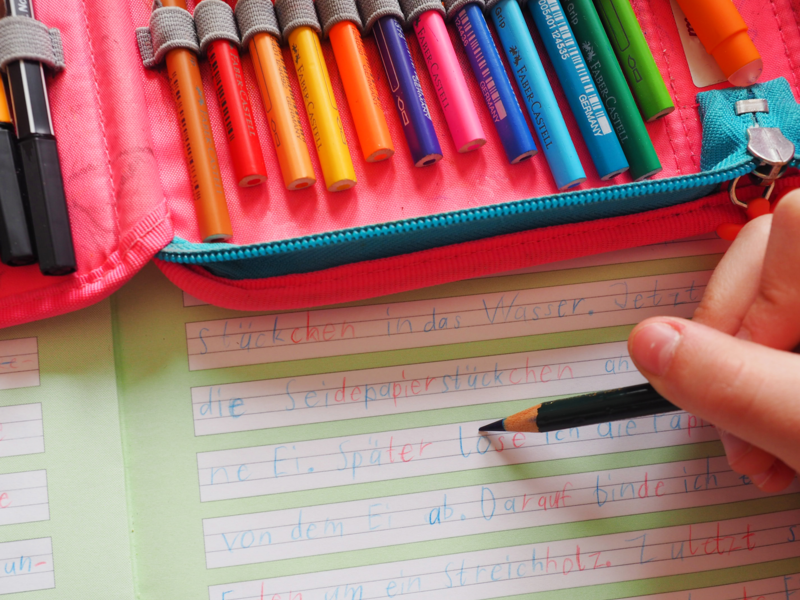 Eine Kinderhand hält einen Buntstift mit blauer Mine und schreibt in ein Schreibheft mit vorgegebenen Linien in einem weißen Schriftfeld Wörter. Der Rest des Heftes ist hellgrün und auf dem Heft liegt ein offenes Federmäppchen mit bunten Buntstiften.