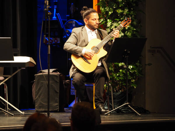 Ein Mann sitzt auf einem Stuhl auf der Bühne, hält und spielt Gitarre. Ein Mikrofon und eine Efeupflanze sind zu sehen.