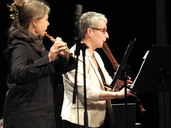 Zwei Frauen spielen Blasinstrumente, die links stehende Querflöte und rechts die Altblockflöte, Mikrofon und Notenständer stehen vor Ihnen.