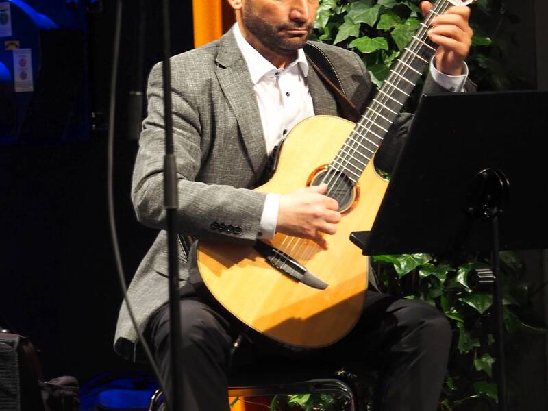 Ein Mann sitzt auf einem Stuhl auf der Bühne mit einer Gitarre und spielt. Im Hintergrund sieht man eine Efeupflanze.