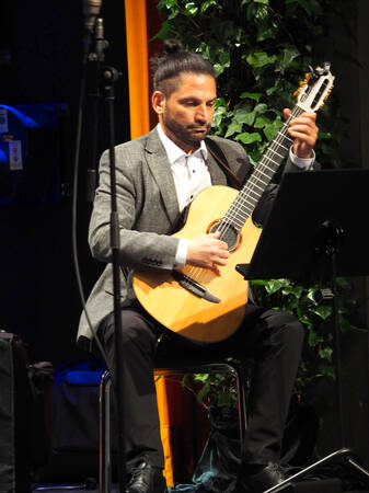 Ein Mann sitzt auf einem Stuhl auf der Bühne mit einer Gitarre und spielt. Im Hintergrund sieht man eine Efeupflanze.