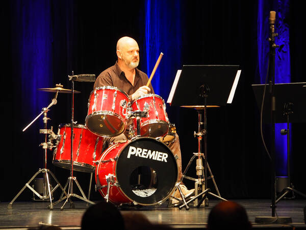 Ein glatzköpfiger Mann sitzt am Schlagzeug, hat zwei Schläger / Drumsticks in der Hand und spielt. Es stehen Notenständer auf der Bühne herum.