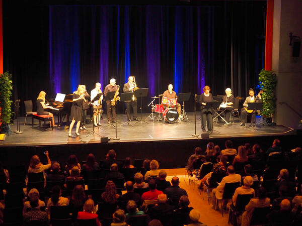 Auf der Bühne stehen 10  Musiker mit verschiedenen Instrumenten, ein großer Flügel ist links zu sehen. Vor der Bühne sieht man das Publikum mit vielen vollen und auch leeren Plätzen.
