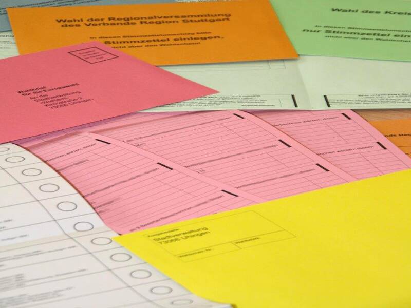 Auf einem Stapel liegen bunte Stimmzettel zur Kommunalwahl aufeinander. Sie sind rot, orange, gelb und grün.