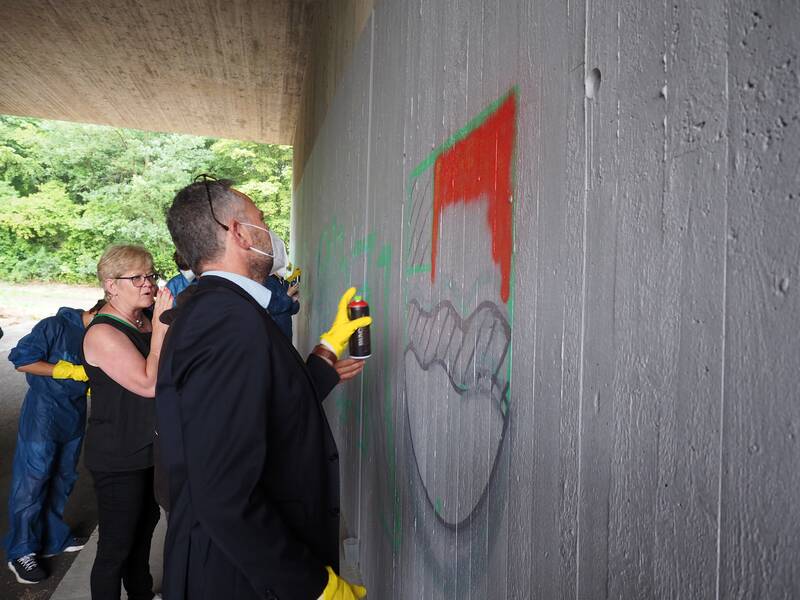 Menschen stehen vor einer Betonwand einer Unterführung. Der Bürgermeister trägt zu seinem Anzug Gummihandschuhe und hat einen Atemschutz vor Nase und Mund, da er mit einer Spraydose auf der legalen Graffiti-Wand rote Farbe aufsprüht.