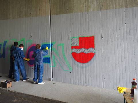 Vor einer grauen Betonwand in einer Unterführung stehen Jugendliche in Ganzkörperschutzanzügen und sprühen auf die legale Graffiti-Wand mit Spraydosen den Schriftzug "Uhingen".