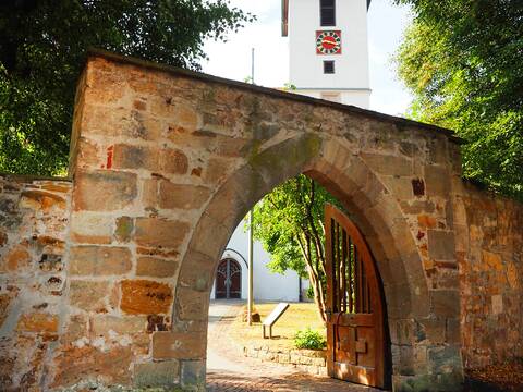 Hinter einem großen Torbogen befindet sich der Innenhof der Cäcilienkirche. Der weiße Kirchturm mit rotem Ziffernblatt und goldfarbenen Zeigern der Kirchturmuhr ragt über die Mauer.