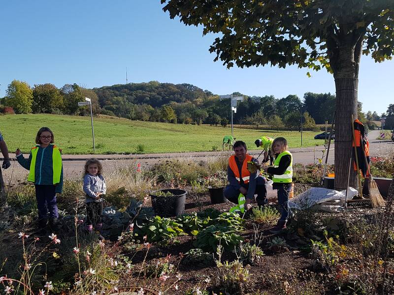 Mehrere Menschen, darunter Kinder und Erwachsene, stehen zum Teil mit neonfarbenen Warnwesten in einem Kreisverkehr, auf dem ein Baum steht. Die Menschen jäten Unkraut, das zwischen einzelnen Pflanzen steht und werfen die Reste in schwarze Eimer. Am Baum lehnen Geräte wie Harke und Besen. Im Hintergrund sind eine Wiese mit angrenzendem Wald und Wohnhäuser zu sehen.