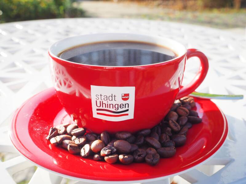 Das Foto zeigt eine rote Kaffeetasse mit Untertasse auf einem weißen Metalltisch. Auf der Tasse ist das Logo der Stadt Uhingen zu sehen. Auf die Untertassen sind einige Kaffeebohne gestreut. 