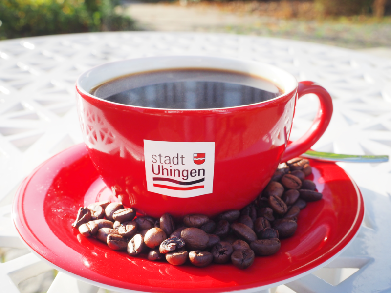 Das Foto zeigt eine rote Kaffeetasse mit Untertasse auf einem weißen Metalltisch. Auf der Tasse ist das Logo der Stadt Uhingen zu sehen. Auf die Untertassen sind einige Kaffeebohne gestreut. 