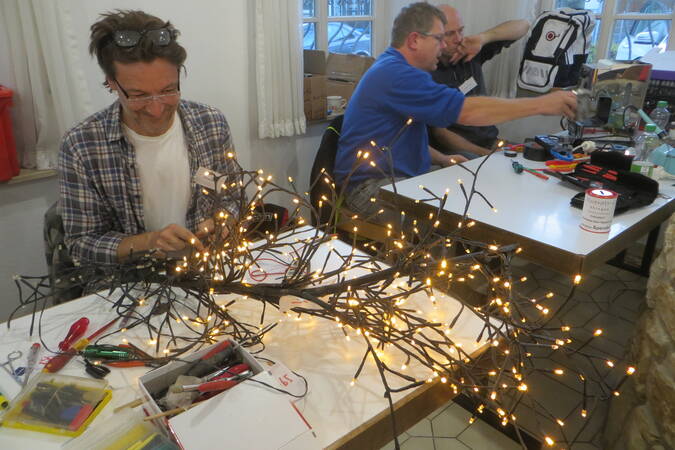 Ein Mann repariert schmunzelnd eine Weihnachtsbeleuchtung mit unzählig vielen kleinen Glühbirnen.  Zwei weitere Männer reparieren zusammen an einer Kaffeemaschine.