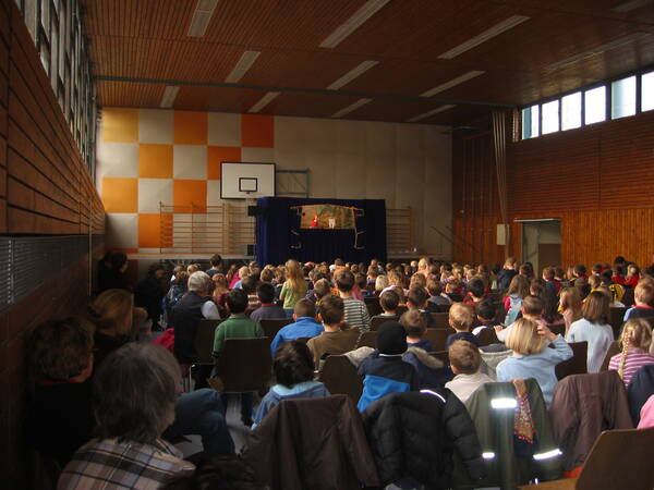 Eine große Halle mit vollbesetzten Stuhlreihen. Man sieht den Rücken, der dort sitzenden Kinder. Sie schauen gespannt auf das Puppenspiel, das im Vordergrund aufgebaut ist. 