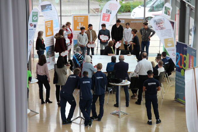 Etwa 30 Personen unterschiedlichen Alters stehen um eine Insel mit Tischen oder um Stehtische vor der Glasfront im Foyer des Uditoriums. Sie sind bei einem Workshop. Es stehen sechs Beachflags und eine Pinnwand um die Tische, die die 17 UN-Ziele zeigen.  