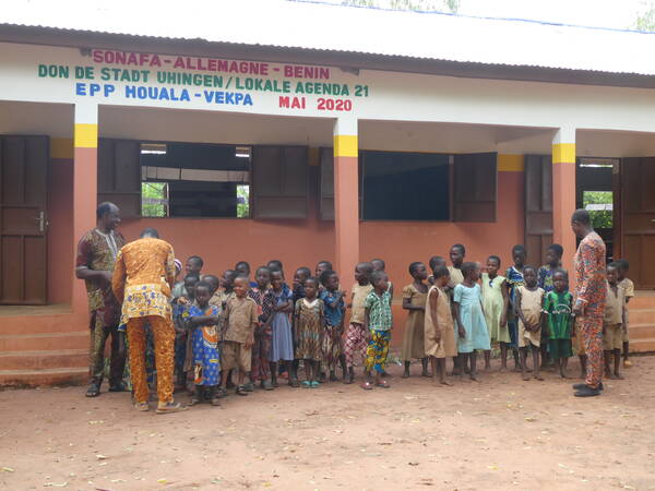 Die Schülerinnen und Schüler der Grundschule in Vekpa stehen mit drei erwachsenen Personen vor dem Schulgebäude. Die Kinder und die Erwachsenen lachen und freuen sich. Auf dem Gebäude ist die Aufschrift  Sonafa Allemagne Benin, don de Stadt Uhingen/Lokale Agenda 21 Mai 2020 zu sehen. 