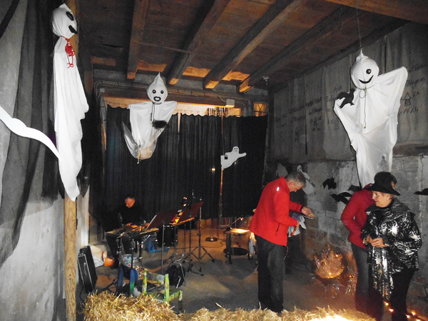 In einer Scheune hängen kleine Gespenster von der Decke. Ein Mann sitzt an einem Schlagzeug. Es gibt Strohballen als Sitzgelegenheiten. Zwei Herren bauen weitere Dekorationsstücke auf. Eine Dame trägt ein Harry-Potter-Kostüm mit glitzerndem  Umhang.