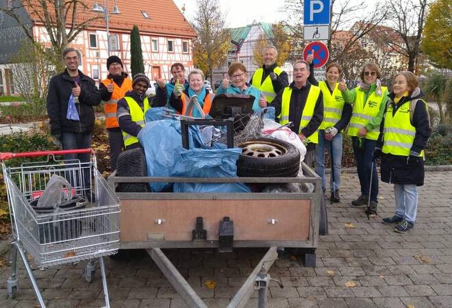 Mehr als 10 Männer und Frauen stehen auf einem großen Platz um einen Anhänger, der mit Müllsäcken gefüllt ist. Die Leute schauen auf die Kamera, lächeln und zeigen den dicken Daumen.  Sie tragen Warnweste und haben augenscheinlich Müll gesammelt. Auch ein alter Einkaufswagen mit einem Eimer darin und ein Autoreifen sind dabei. 