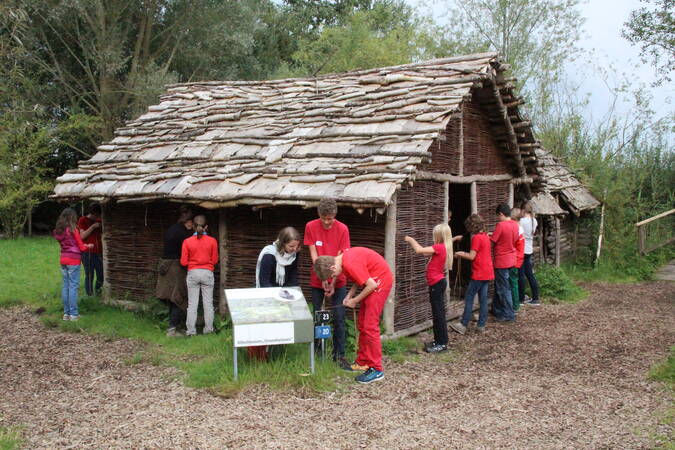Über 10 Jugendlichen stehen rund um eine steinzeitliche Hütte und binden Holzstöckchen zusammen. 