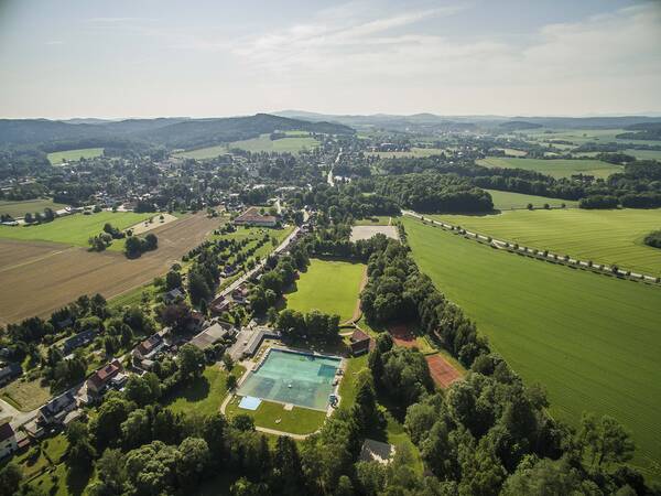 Oppach inmitten von grünen Wiesen und braunen Ackerflächen sowie Wäldern im Sommer von der Luft aus betrachtet. Im Vordergrund ist ein Freibad, dessen Wasser türkis schimmert, inmitten von Bäumen. Die daneben befindliche Straße, an der sich Häuser und Bäume abwechseln, zieht sich fast bis zum Horizont. Auffällig ist, dass sich viele hohe Bäume im Gemeindegebiet befinden. Am Horizont sind Hügel und Schleierwolken am blauen Himmel.