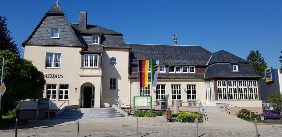 Das Rathaus in Oppach im Sommer mit dem Vorplatz aus Pflastersteinen Die Fassade ist grau, die Fenster weiß mit weißem Fensterkreuz. Das Dach ist dunkel gedeckt. Vor dem Rathaus wehen die Flaggen von Deutschland (senkrechte Streifen in schwarz, rot und gelb) sowie von Sachsen (senkrechte Streifen grün und weiß sowie Sachsens Wappen).