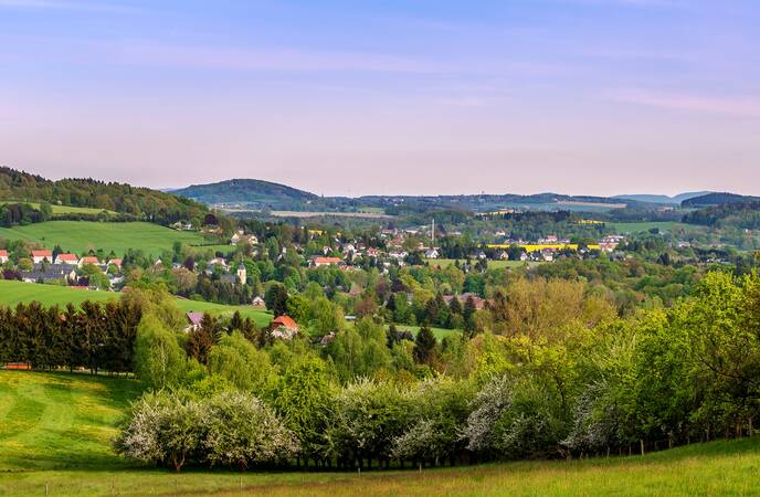 Luftaufnahme von Oppach: Im Vordergrund ein Wald und dahinter grüne Wiesen sowie Häuser verstreut zwischen Bäumen, gelben Rapsfeldern und die hügelige Landschaft zeichnet sich am Horizont vor dem blauen Himmel mit wenigen Wolkenschleiern ab.