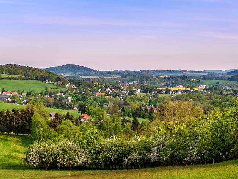 Luftaufnahme von Oppach: Im Vordergrund ein Wald und dahinter grüne Wiesen sowie Häuser verstreut zwischen Bäumen, gelben Rapsfeldern und die hügelige Landschaft zeichnet sich am Horizont vor dem blauen Himmel mit wenigen Wolkenschleiern ab.
