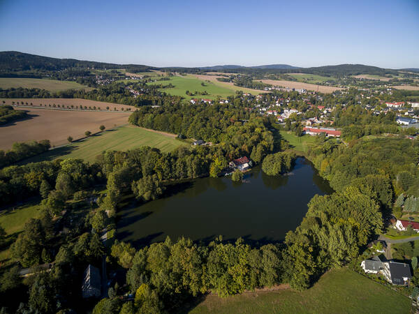 Oppach von der Luft aus gesehen: Im Vordergrund ist der Gondelteich, dessen Wasser dunkel schimmert. Der Teich ist von Bäumen umrandet und ein Haus befindet sich am See. Dahinter wechseln sich Häuser, Bäume, Felder und Wälder ab. Eine leichte Hügelkette ist am Horizont, der Himmel ist blau.