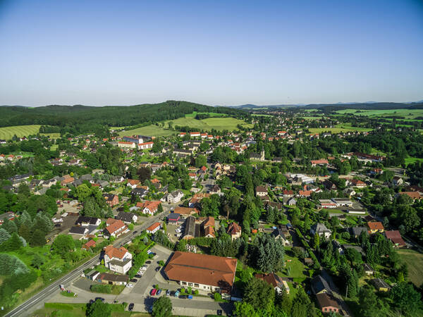 Luftaufnahme von Oppach, Partnergemeinde von Uhingen: Dächer von großen und kleinen Gebäuden sind inmitten vieler Bäume, auf einer Straße fahren Fahrzeuge am Horizont sind grüne Felder und Wälder sowie der blaue Himmel.