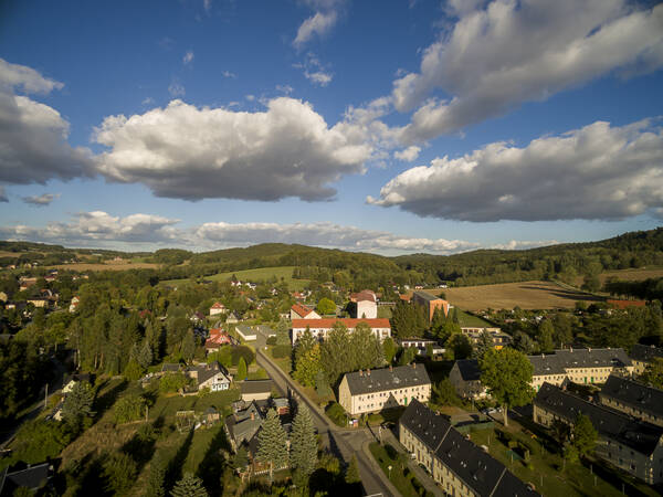 Während am blauen Himmel größere Wolken vorbeizeihen liegt am Boden die Gemeinde Oppach. An einer Straße befinden sich mehrere zweigeschossige Mehrfamilienhäuser mit dunklen Dächern in einer Neubausiedlung sowie vereinzelt Häuser umgeben von vielen Bäumen. Die hügelige Landschaft ist von Wäldern und Feldern durchzogen.