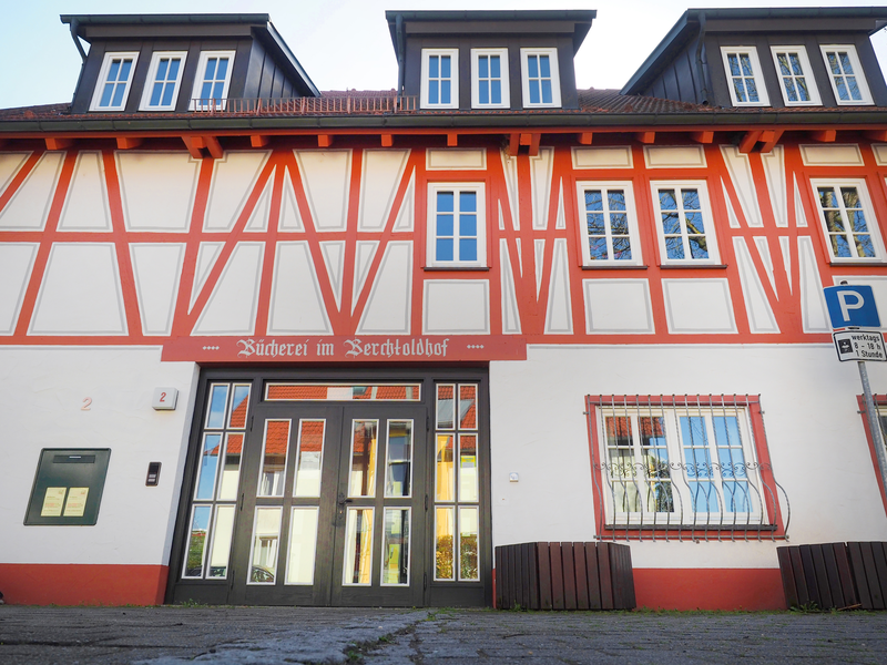 Die Außenansicht der Stadtbücherei: ein weißes Gebäude im Fachwerkstil mit roten  Balken und einer breiten Glastür