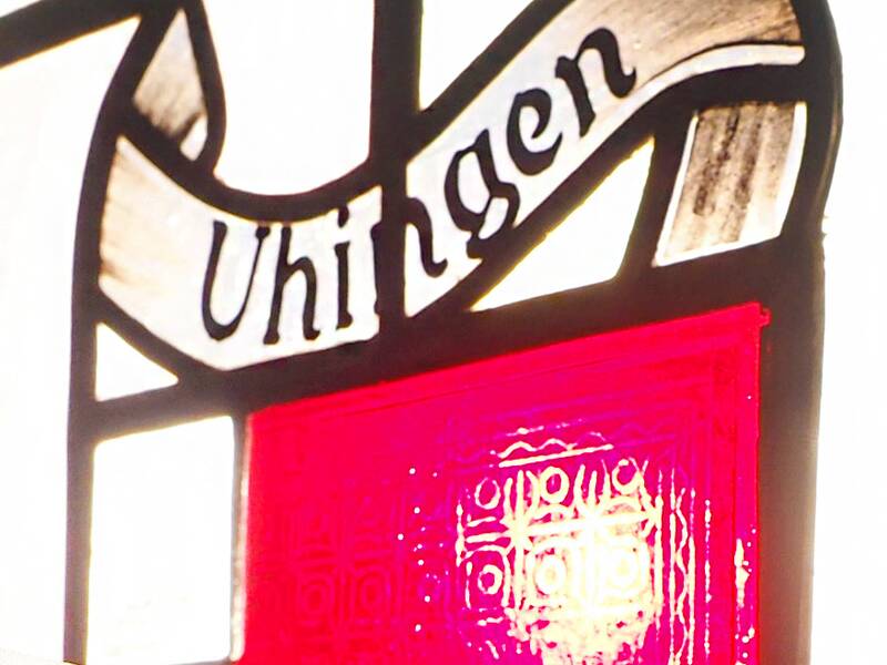  Auf dem Fenster aus Buntglas ist das Wappen der Stadt Uhingen zur Hälfte zu sehen. Links oben ist ein weißes Viereck auf rotem Grund. Das Wappen wird von der Sonne angeleuchtet.