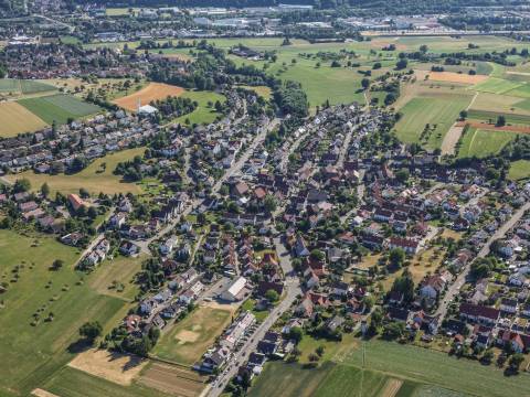 Luftaufnahme von Holzhausen im Sommer. Rings um die Häuser sind grüne Bäume und gelbe Felder zu sehen.