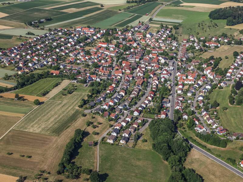 Luftaufnahme von Holzhausen im Sommer. Rings um die Häuser sind grüne Bäume und gelbe Felder zu sehen.