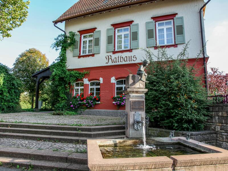 Das Rathaus in Holzhausen: Das Erdgeschoss ist rot, die oberen Geschosse beige. An den Fenstern blühen Geranien. Vor dem Rathaus plätschert ein Brunnen.