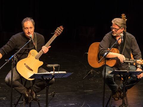 Zwei sitzende Männer mit jeweils einer Gitarre in der Hand und einem Mikrofon zum Singen.