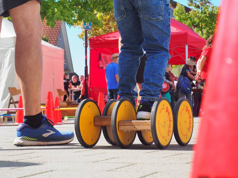 Die Beine eines Kindes stehen auf einem Pedalo: ein Koordinationstrainer mit sechs großen Rädern und die beiden Trittflächen senken und heben sich mit jedem Schritt des Nutzers, sodass sich das Pedalo vorwärts bewegt. Das Pedalo ist Teil einer Spielstraße, bei der Vereine an Ständen sich und ihre Aktivitäten präsentieren. Im Vordergrund ist ein roter Verkehrskegel und dahinter sind Menschen an Ständen.