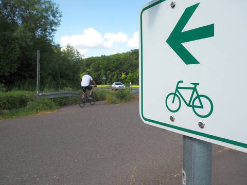 Auf einem Radweg radelt ein Mann neben Bäumen und einer befahrenen Straße. Rechts im Vordergrund ist ein Schild mit dem Hinweis auf ein Fahrradschild.
