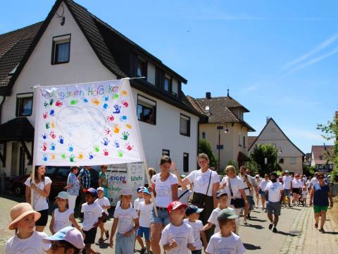 Bei einem Festumzug im Sommer gehen Kinder in weißen Shirts und mit Mütze, erwachsene Frauen tragen ein weißes Lacken als Banner, auf dem bunte Handabdrücke der Kinder sind