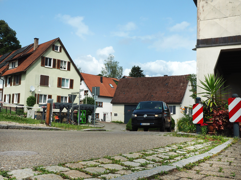 Ein Auto fährt durch eine Engstelle einer Ortsdurchfahrt, an deren Seiten Häuser und eine Rasenfläche sind.