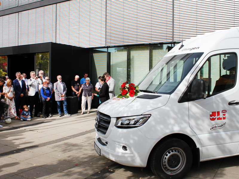 Mehrere Menschen stehen vor einem Gebäude mit Glasfassade und schauen auf einen weißen Kleinbus, auf dessen Motorhaube ein Blumenstrauß mit roten Blumen liegt.