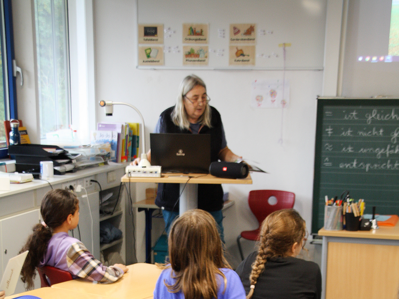 Kinder sitzen in einem Klassenzimmer immer zu viert an kleinen Tischinseln, an einem Rednerpult steht eine Frau und liest eine Geschichte vor.