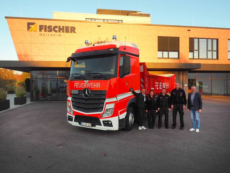 Vor einem modernen Gebäude mit der Aufschrift "Fischer Weilheim" steht ein großer roter Feuerwehr-Lastwagen, umgeben von fünf Personen  (von rechts): Bürgermeister Matthias Wittlinger, Kommandant Steffen Kwiatkowski, Gerätewart Heiko Streicher sowie Verwaltungsmitarbeiterin und FFW-Mitglied Jenny Streicher sowie FISCHER-Geschäftsführer Frank Bahner.		