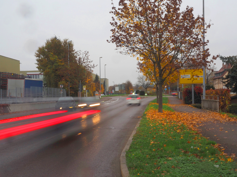 Eine Straße bei schlechtem Wetter zeigt Autos in Bewegung mit verwischten Rücklichtern, Herbstbäume und ein Verkehrszeichen am Rand.