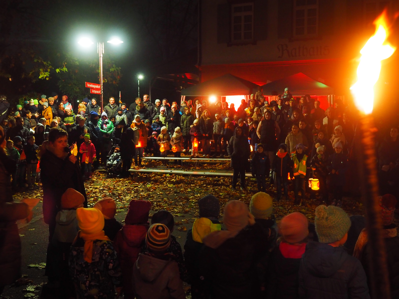 Viele Menschen, darunter viele Kinder mit bunten Mützen und Laternen, versammelen sich in der Dunkelheit vor dem Rathaus im Uhinger Stadtteil Holzhausen zum Laternenumzug und Martinsspiel. Damit feiert der Kindergarten Hofäcker sein 30-jähriges Bestehen. 