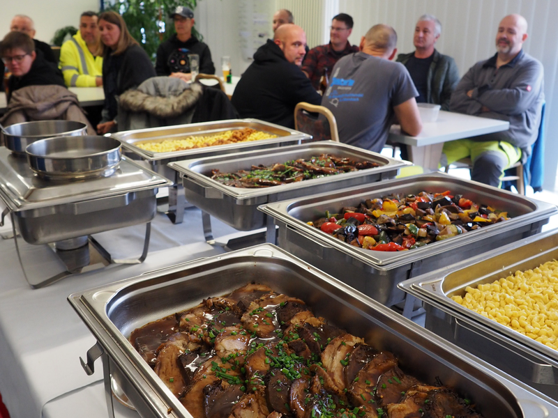 Ein Buffet mit verschiedenen warmen Speisen steht bereit, während mehrere Personen von Bauhof, Kläranlage und Hausmeister-Team der Hieberschule an Tischen im Hintergrund sitzen.