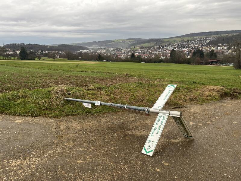 Durch stumpfe Gewalteinwirkung wurde ein Schild aus der Verankerung gerissen. Es liegt auf einem Feldweg neben einem Feld, im Hintergrund befindet sich Uhingen.