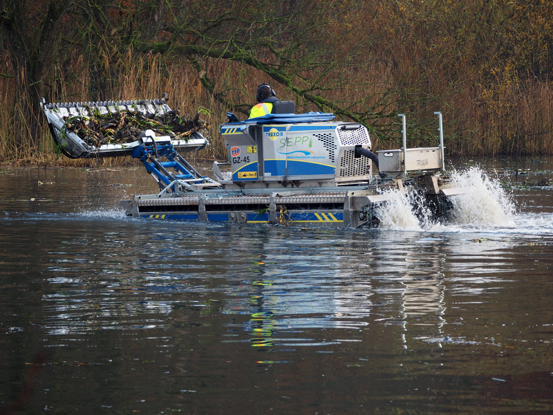 Ein spezielles Amphibienboot schwimmt auf dem Charlottensee und entfernt mit einer großen Schaufel Wasserpflanzen, Schilf und Teichmummel, aus dem Wasser.