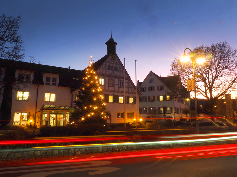 Ein erleuchteter Weihnachtsbaum steht vor dem Uhinger Rathaus in der Dämmerung, während Lichter von vorbeifahrenden Autos Streifen im Vordergrund hinterlassen.