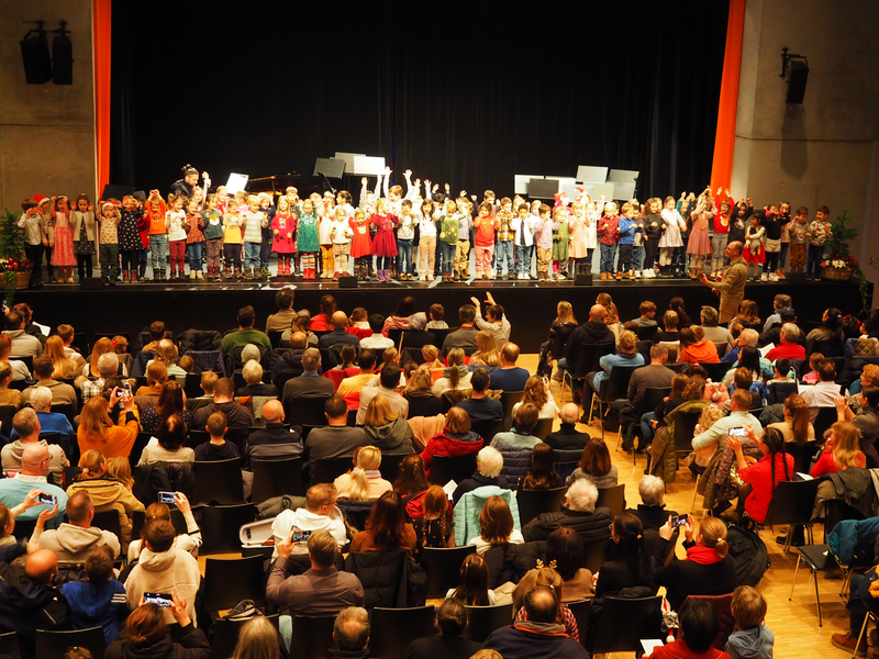 Eine Gruppe von Kindern steht auf der Bühne des Uditoriums und singt vor einem Publikum, das im großen Saal sitzt.