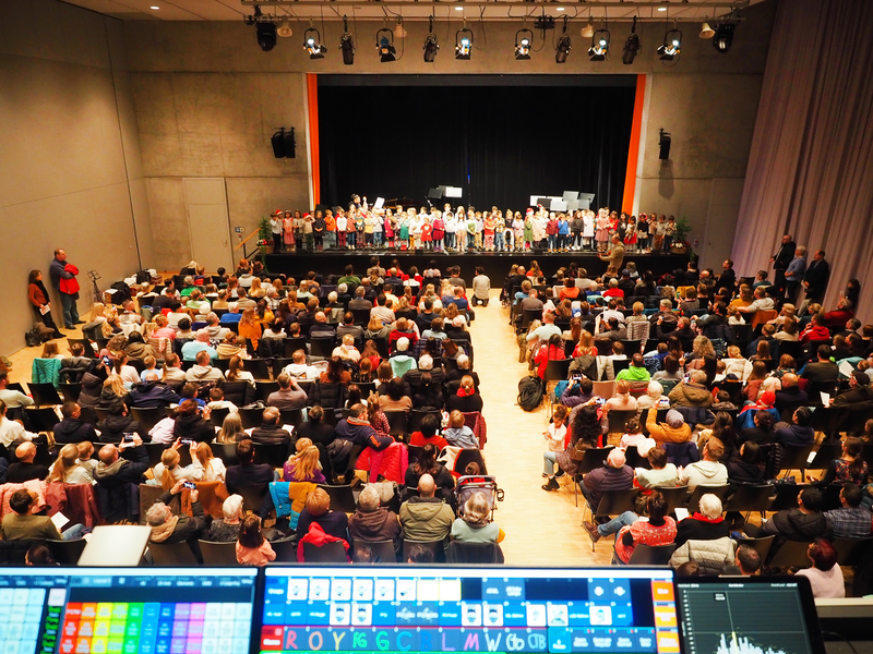 Ein voller Saal mit Zuschauern blickt auf die Bühne im Uditorium, auf der eine große Gruppe von Kindern steht.